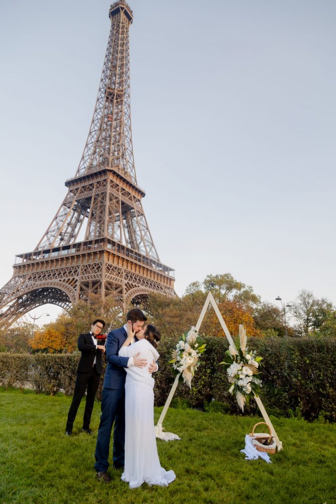 Casal de noivos se beijando em frente à Torre Eiffel durante um elopement romântico com decoração floral e violinista ao fundo
