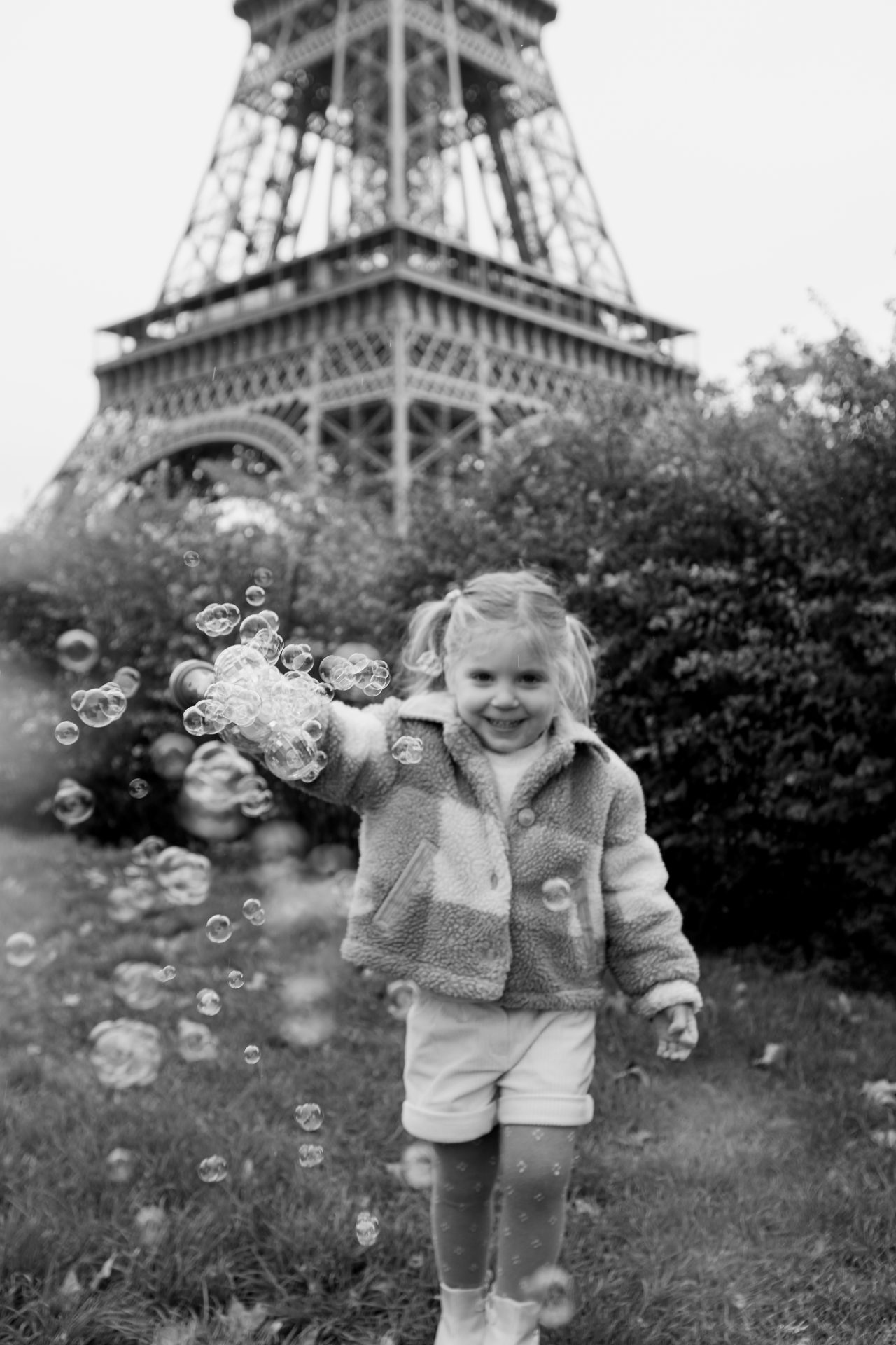 Daughter smiling at camera blowing bubbles in flower garden in front of Eiffel Tower during Paris photoshoot