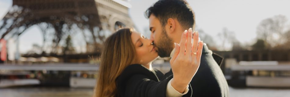 Woman showing her engagement ring while kissing her fiancé in front of the Eiffel Tower after a romantic proposal in Paris.