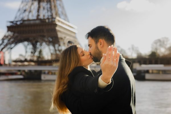 Woman showing her engagement ring while kissing her fiancé in front of the Eiffel Tower after a romantic proposal in Paris.
